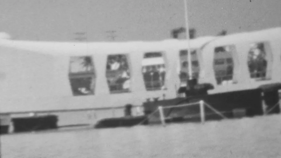 1962 black-and-white 8mm frame showing the curved Arizona Memorial at Pearl Harbor across the water, with open window bays, railings, and small visitor figures near the structure.
