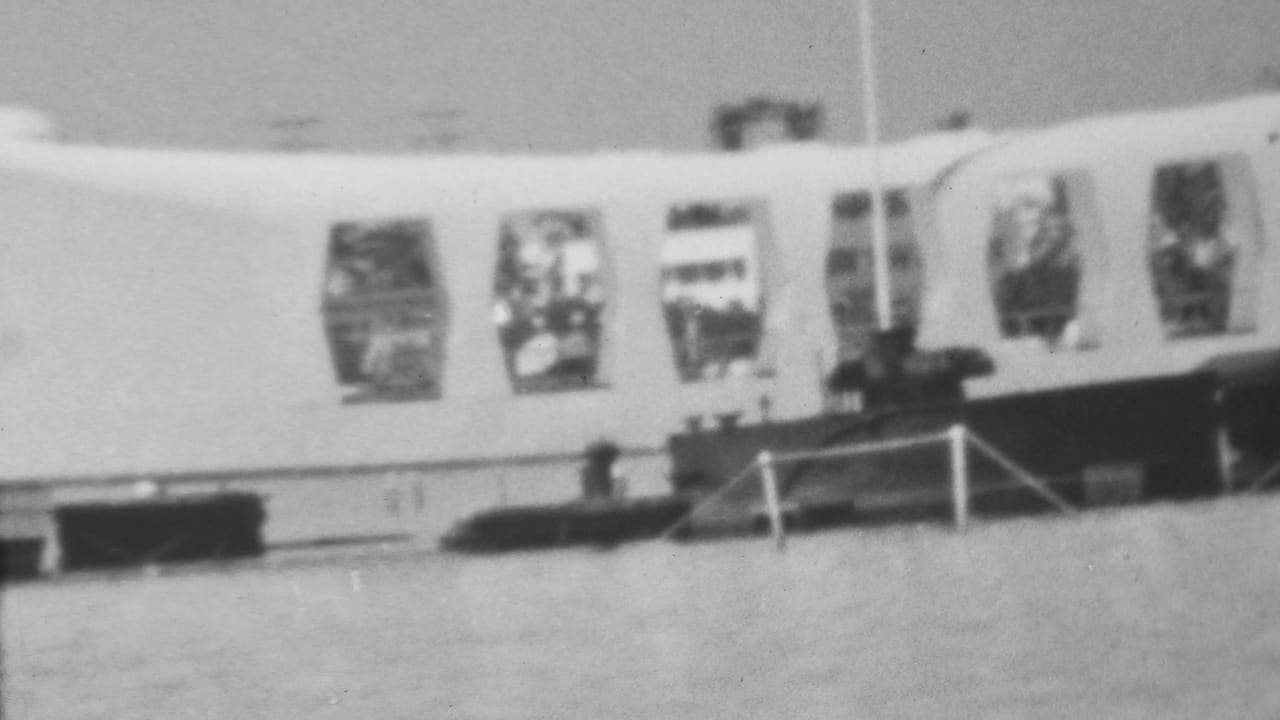1962 Pearl Harbor frame showing a closer section of the Arizona Memorial facade, with repeated window openings, waterfront railing, and small visitors visible near the base.