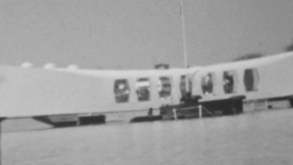 Wide 1962 home-movie frame of the Arizona Memorial at Pearl Harbor, with water in the foreground, the curved white structure, window bays, and small visitors along the railing area.