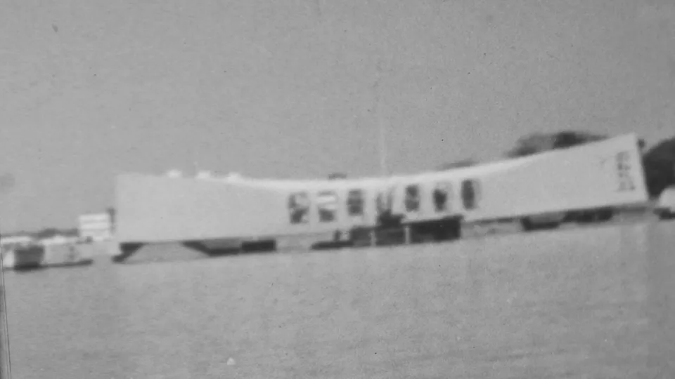 Distant 1962 black-and-white 8mm frame showing the Arizona Memorial at Pearl Harbor stretched across the water, with small window openings and a few indistinct figures near the structure.