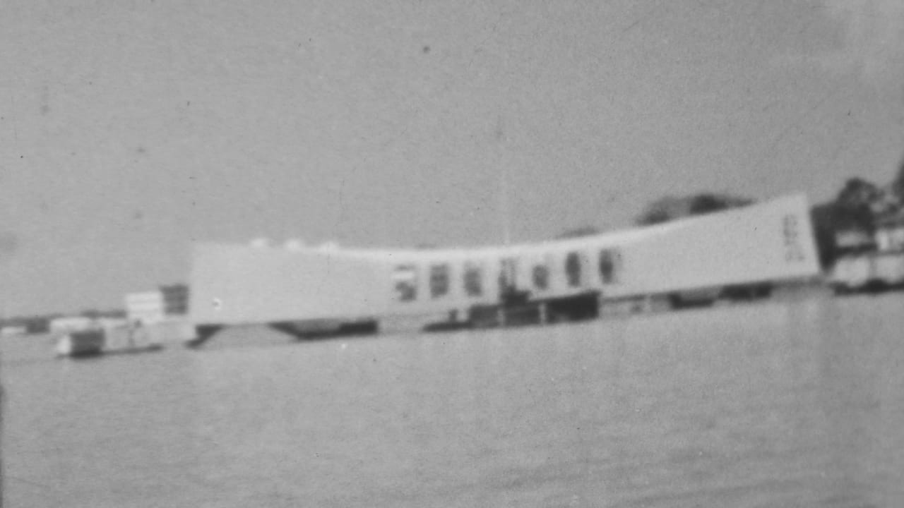 Black-and-white home-movie frame of the long white Pearl Harbor memorial form over water in Honolulu, seen with soft focus and motion blur.