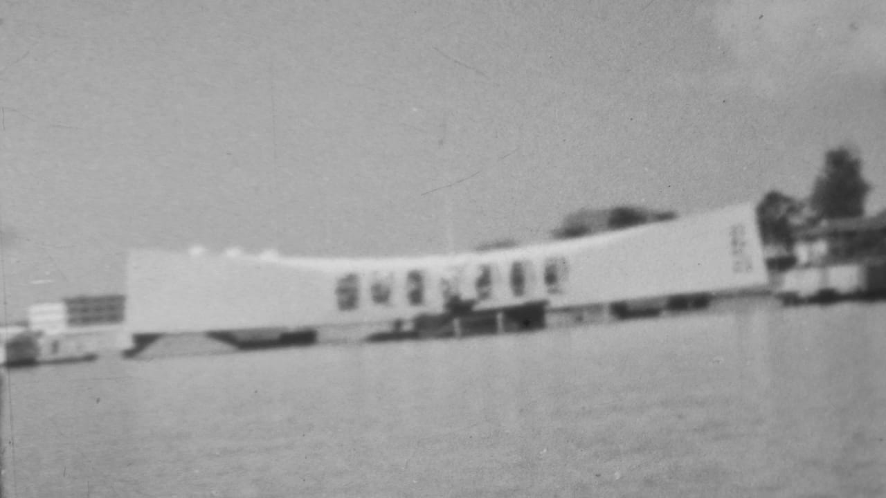 1962 black-and-white 8mm frame from Honolulu showing the Pearl Harbor memorial structure across calm harbor water under a cloudy sky.
