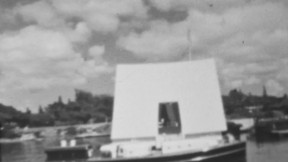 1962 Honolulu home-movie frame showing the white Pearl Harbor memorial from across the harbor, with a soft-focus shoreline and clouds behind it.