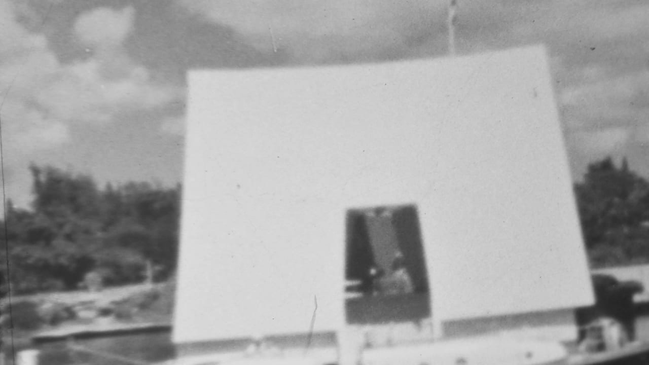 Black-and-white 1962 Pearl Harbor home-movie frame showing an angular concrete structure with a dark entrance, tiny visitor silhouettes, trees, and open sky.