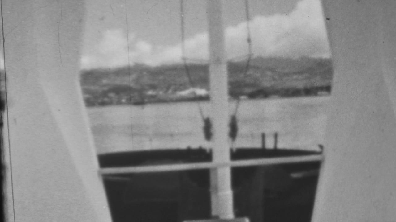 1962 Honolulu waterfront view, likely near Pearl Harbor, framed by a vertical structural opening with calm water, distant land, and cloudy sky beyond.
