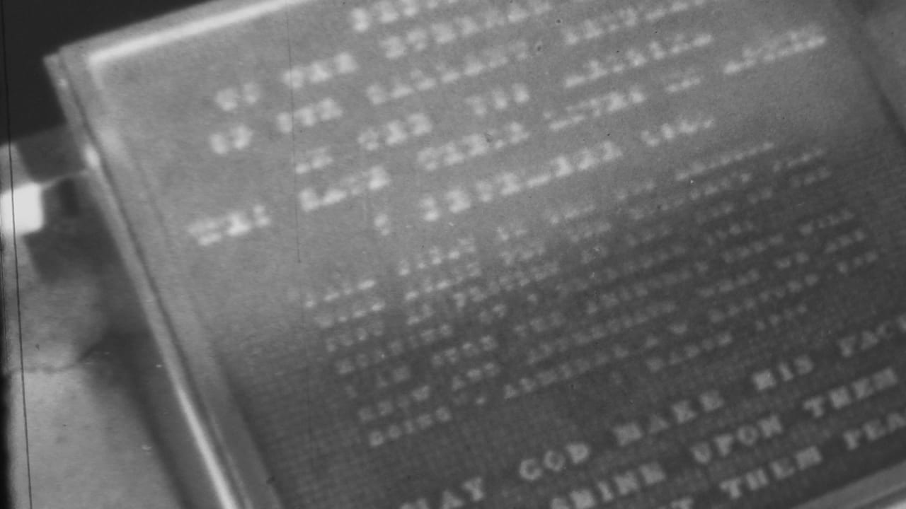 Monochrome visitor-shot frame showing the same engraved plaque with partially legible religious wording and soft 8mm texture.