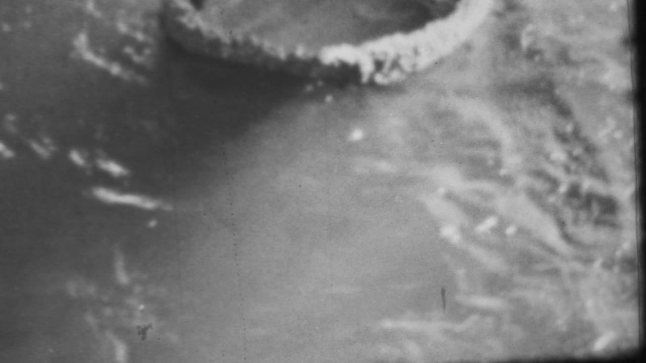 1962 Honolulu-attributed 8mm frame showing a blurred close view of shallow water and pale shoreline texture, with dark wet rock at the edge.