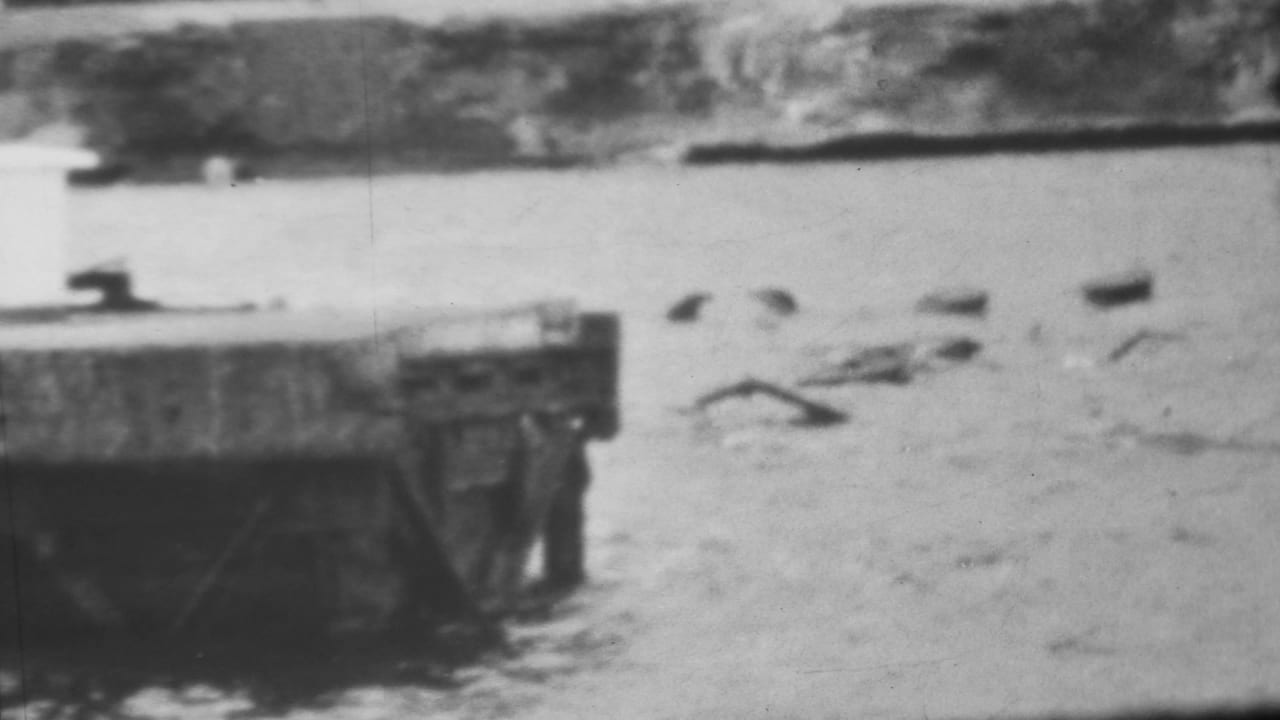 Black-and-white 1962 Pearl Harbor-area 8mm frame showing a concrete pier or broken bridge edge beside turbulent water and floating debris near a low shoreline.