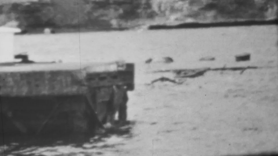 Black-and-white 8mm frame from the 1962 Honolulu footage showing the corner of a concrete pier or bridge remnant with rippling water and debris moving nearby.