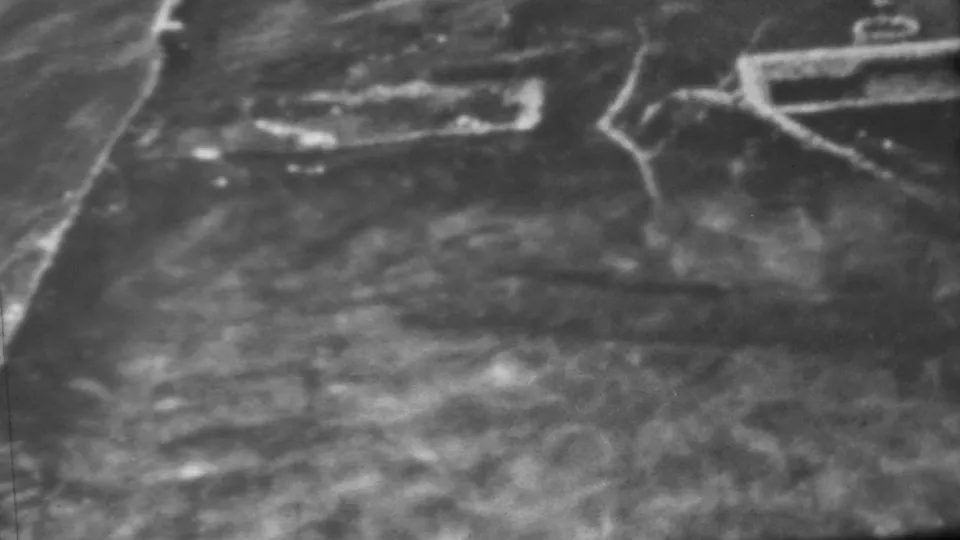 Soft black-and-white 1962 Pearl Harbor-area frame showing a broader shoreline or embankment view with water in the foreground and pale paths or roads crossing the land.