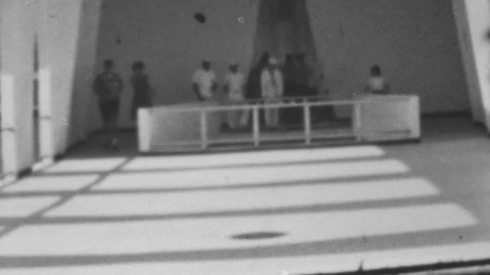 A 1962 Pearl Harbor shipboard frame shows several people, including apparent sailors in light uniforms, standing or walking on a bright deck beside railings and structural supports.