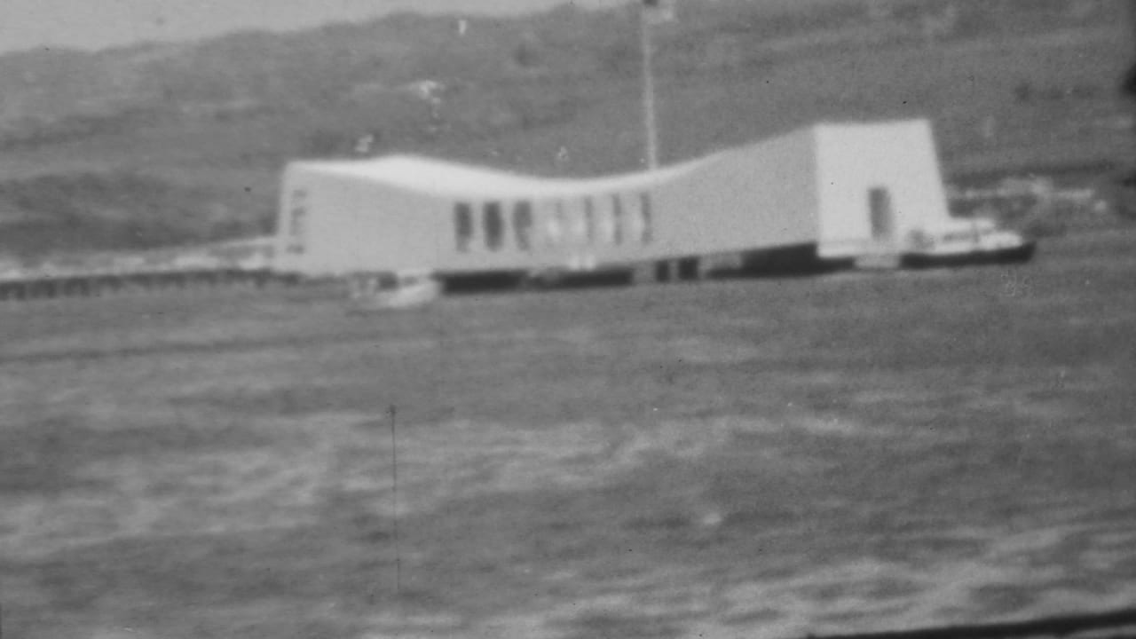 1962 Pearl Harbor home-movie frame showing the white USS Arizona Memorial across the water with a low hillside in the background.