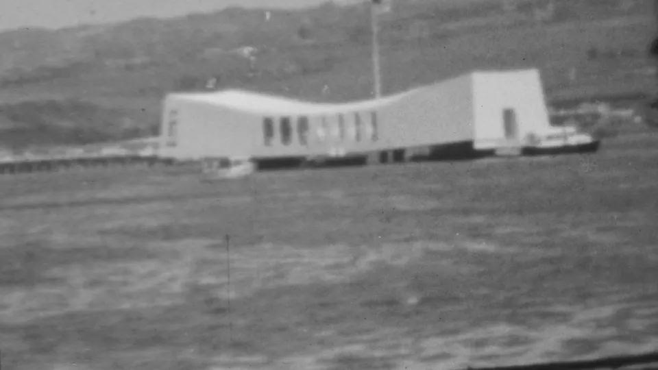 1962 Pearl Harbor home-movie frame showing the white USS Arizona Memorial across the water with a low hillside in the background.