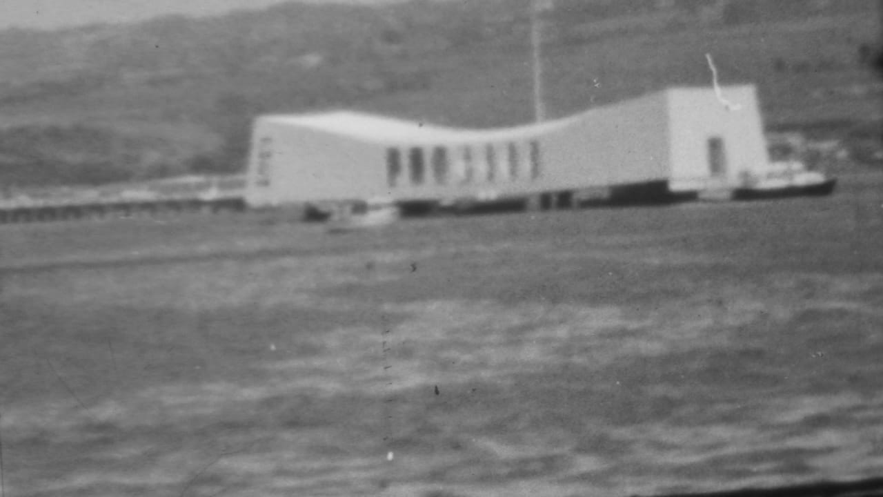 White memorial structure near the water with hillside and shoreline behind it.