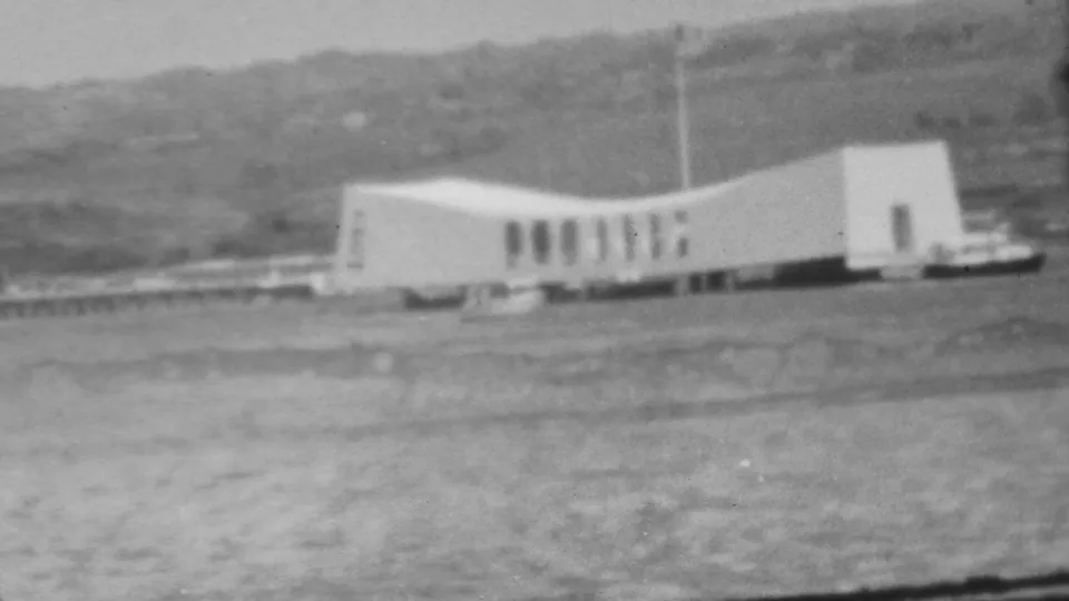 Black-and-white 8mm frame from Pearl Harbor showing the USS Arizona Memorial from a visitor viewpoint across the water.