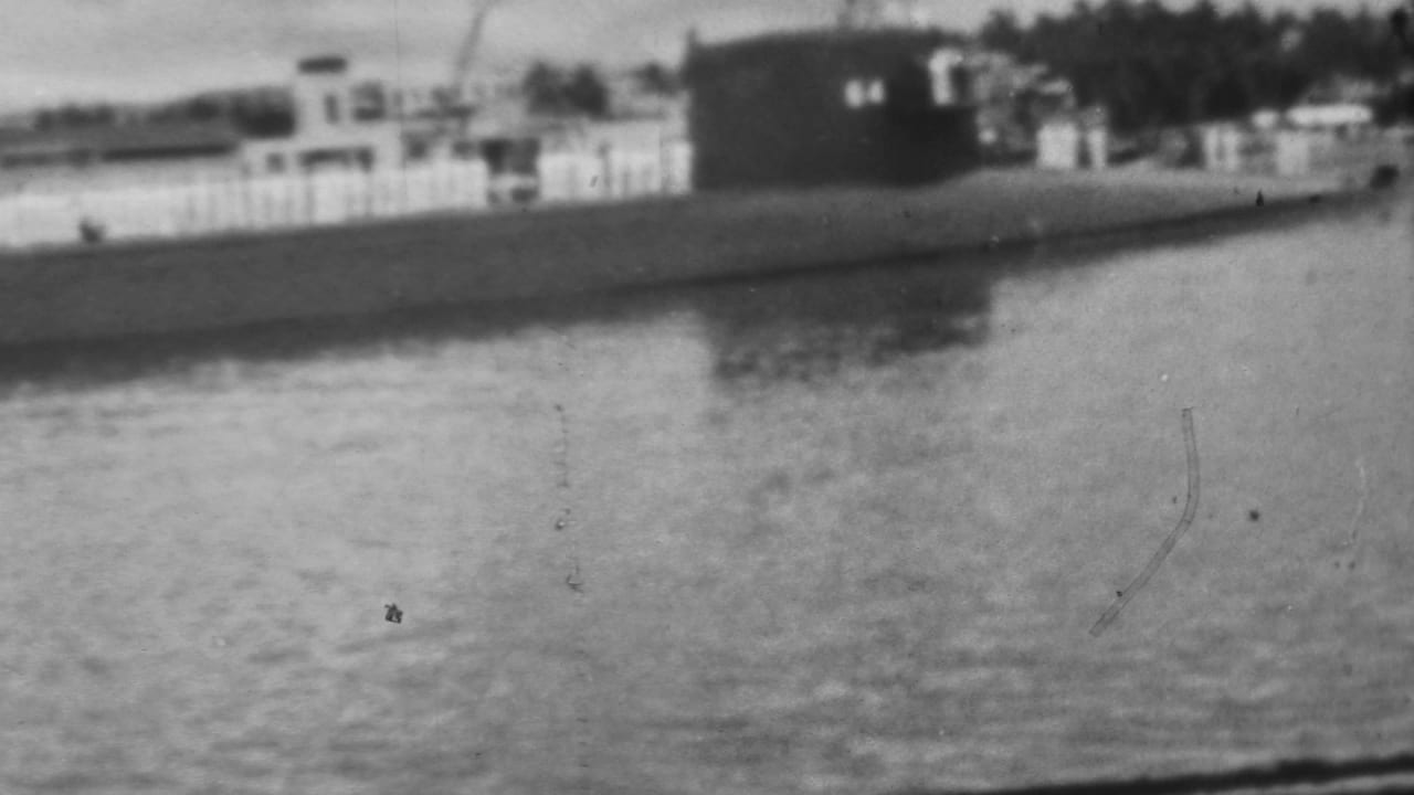 1962 Pearl Harbor footage showing a surfaced submarine viewed from across calm water, with shoreline buildings, fencing, and trees in the background.