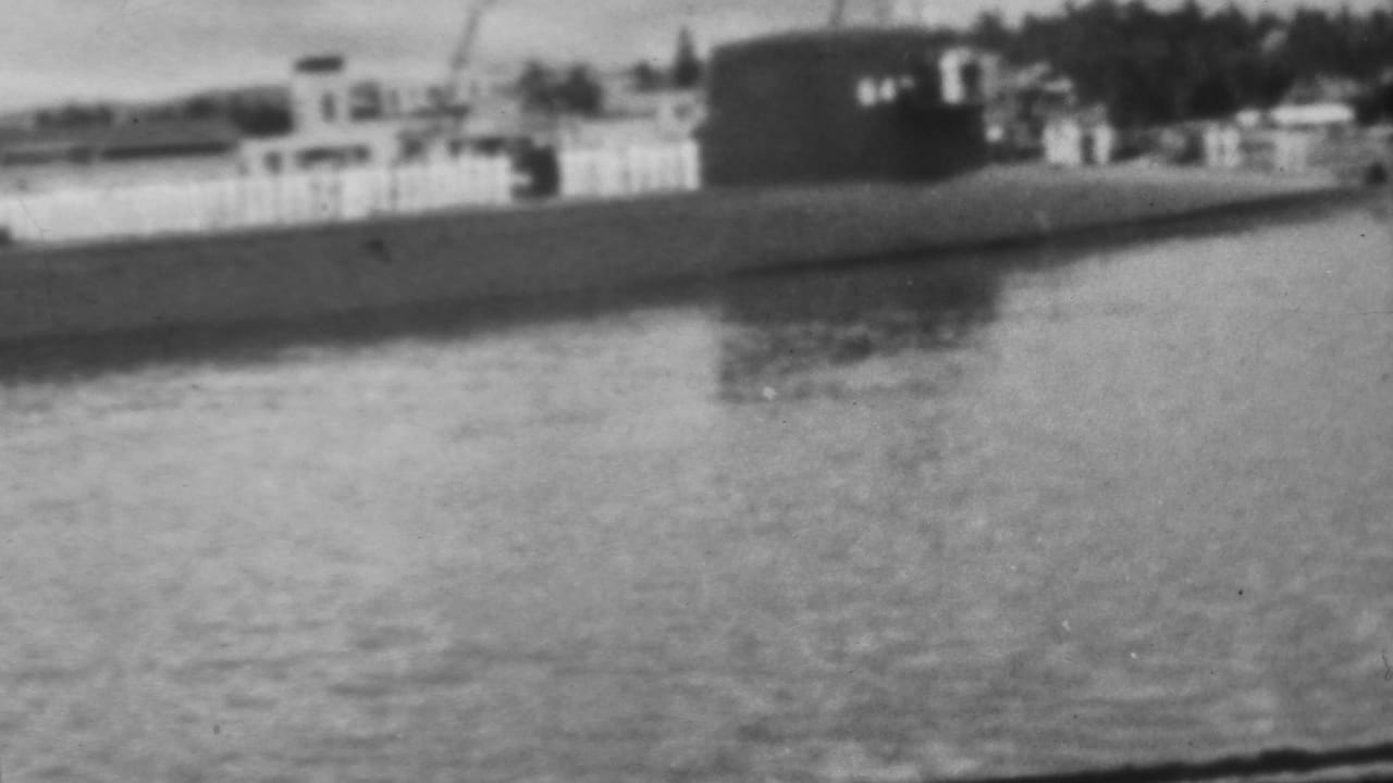 Distant submarine on harbor water with its conning tower near shoreline structures.