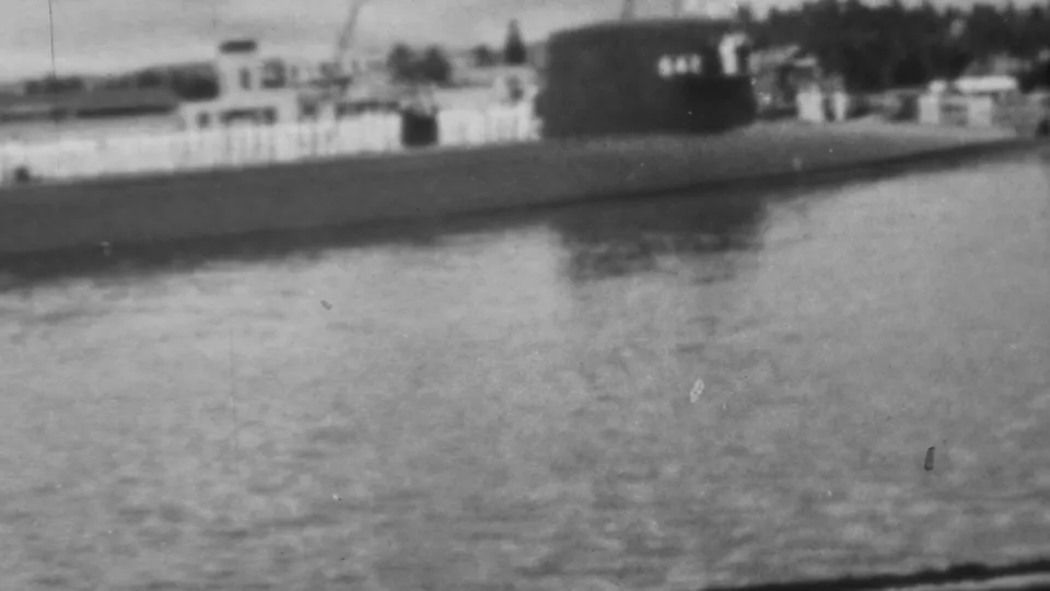 Soft black-and-white 8mm frame of a submarine at Pearl Harbor, with the hull crossing the lower frame and shoreline fencing and buildings behind it.