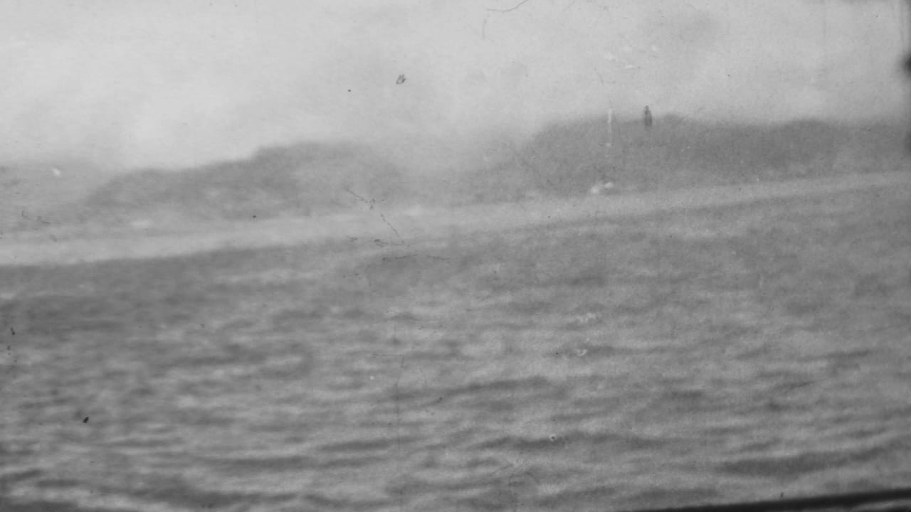 A 1962 8mm view from the water at Pearl Harbor shows choppy harbor surface, hazy hills, and faint shoreline or port structures in the distance.