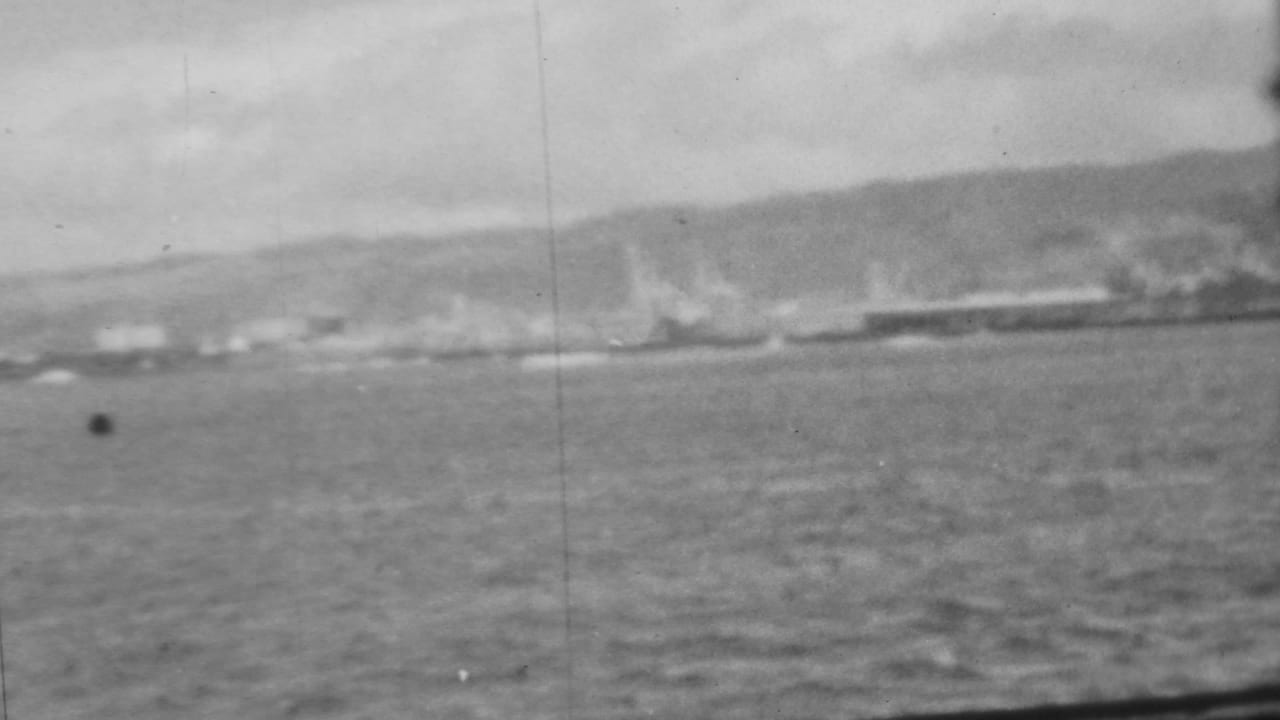 From a moving ship, the frame shows Pearl Harbor water in the foreground with a hazy shoreline, low port forms, and hills beyond.