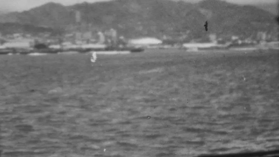 A wider 1962 Pearl Harbor frame shows dark rippled water, a hazy industrial-looking shoreline, pale waterfront structures, and hills in the background.