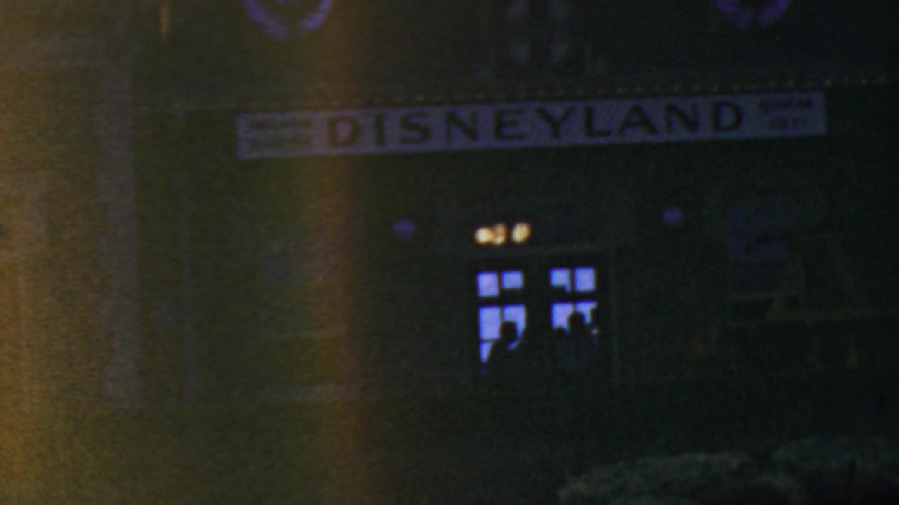 1964 8mm frame from Anaheim showing the Disneyland entrance sign at night above a dark building facade with glowing windows and indistinct visitor silhouettes.