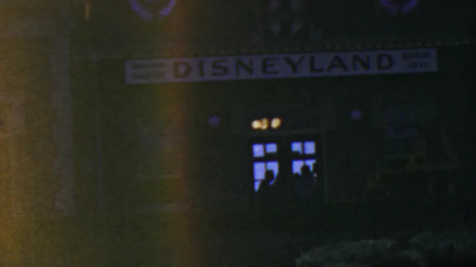 1964 8mm frame from Anaheim showing the Disneyland entrance sign at night above a dark building facade with glowing windows and indistinct visitor silhouettes.