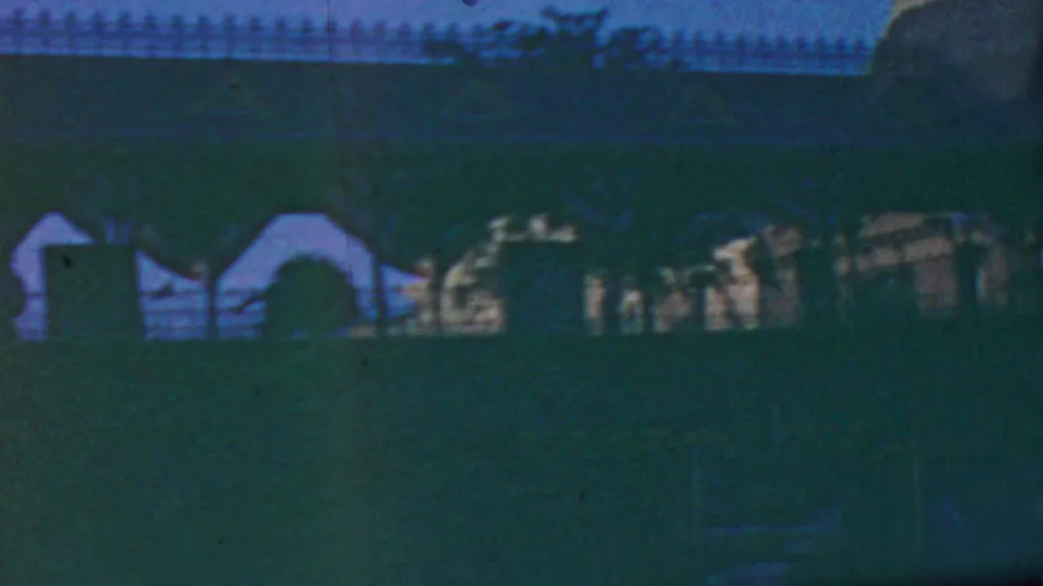 1964 Anaheim home-movie frame with distant park architecture, dark lawn foreground, fence detail, and bright sunlit highlights softened by 8mm grain.