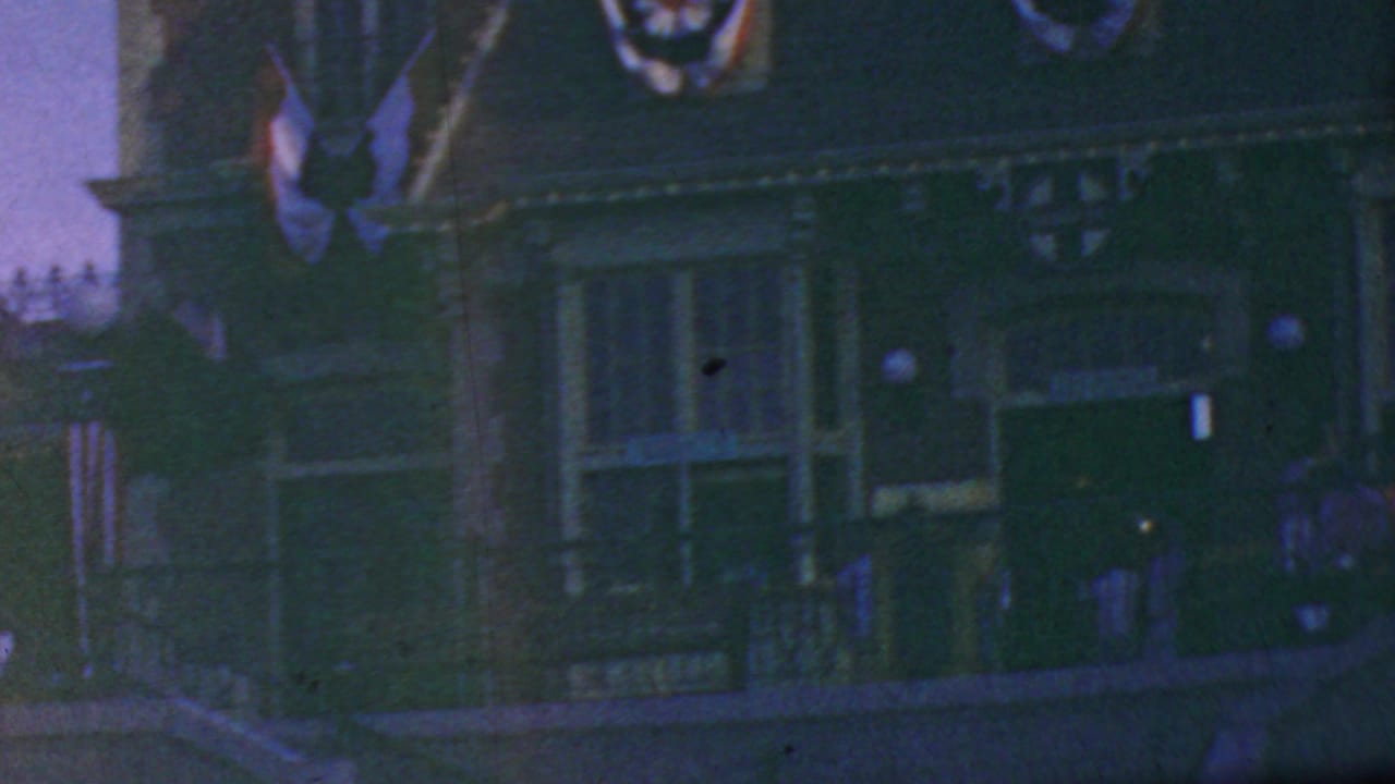 1964 Disneyland home-movie frame showing a dim decorated facade with flags, bunting, railings, lamps, and distant figures along the route.