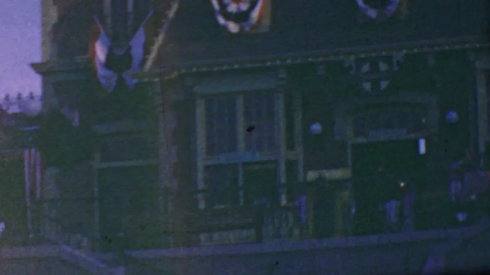 1964 Disneyland home-movie frame showing a dim decorated facade with flags, bunting, railings, lamps, and distant figures along the route.