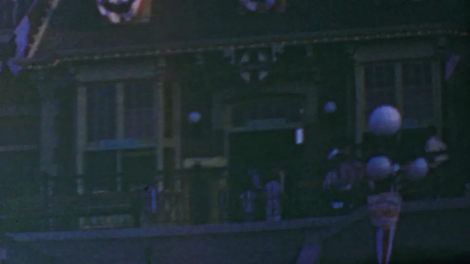 1964 Disneyland nighttime parade-route frame with bunting, globe lamps, railings, and uniformed figures moving near the decorated facade.