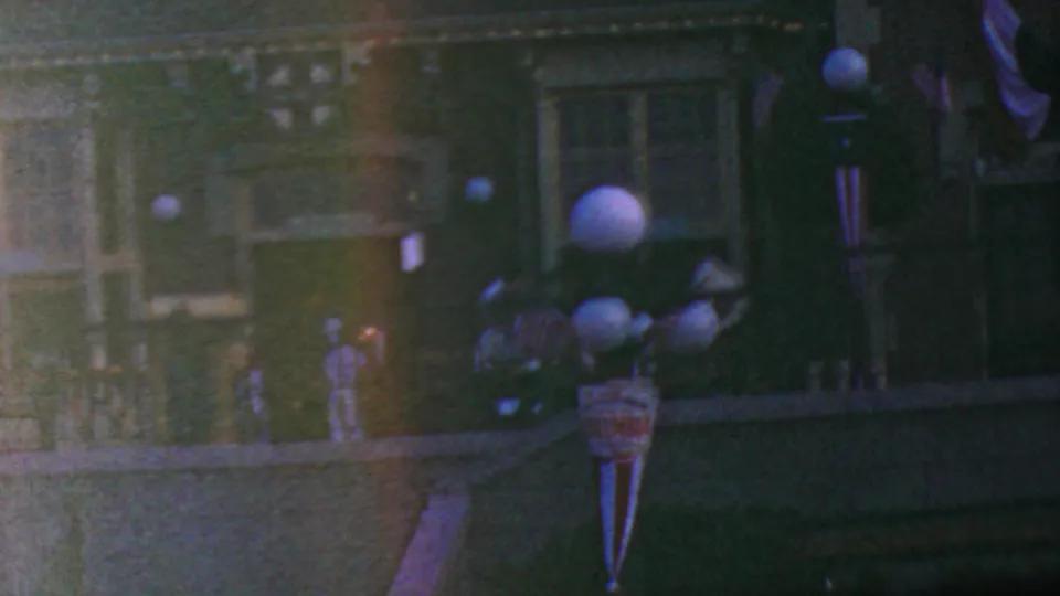 1964 Disneyland home-movie frame showing a closer low-light view of the decorated route with railings, globe lamps, facade details, and a hanging sign or banner.