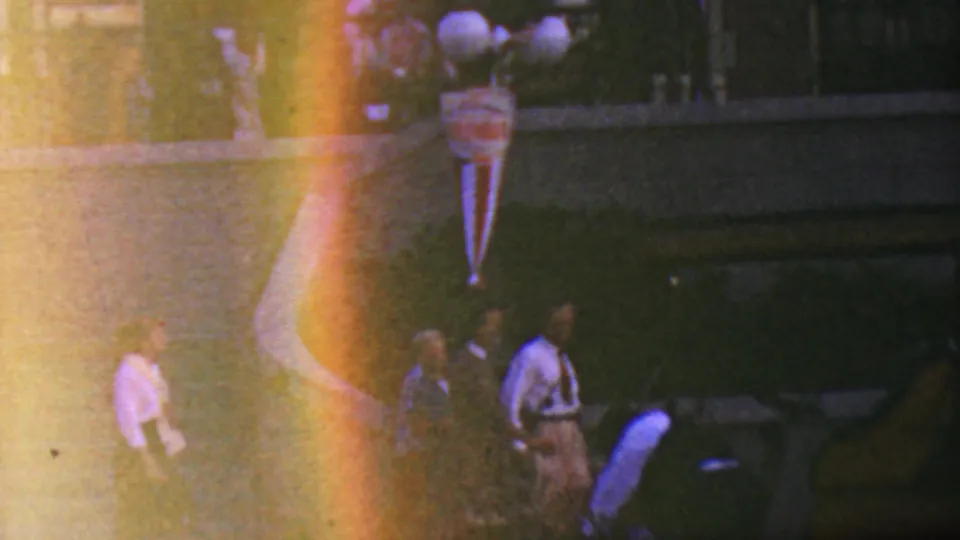 1964 Disneyland nighttime frame with several people walking near the parade route, globe lamps and bunting above them, and bright lens flare from the 8mm camera.
