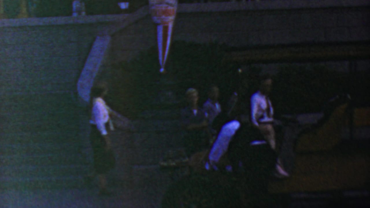 1964 Anaheim Disneyland home-movie frame showing adults in low light near a railing and raised event structure, with heavy grain and color shift.