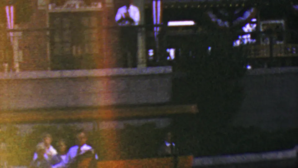 Grainy 1964 Anaheim Disneyland nighttime frame showing a balcony or raised viewing area with a person above and figures below near ceremony staging.