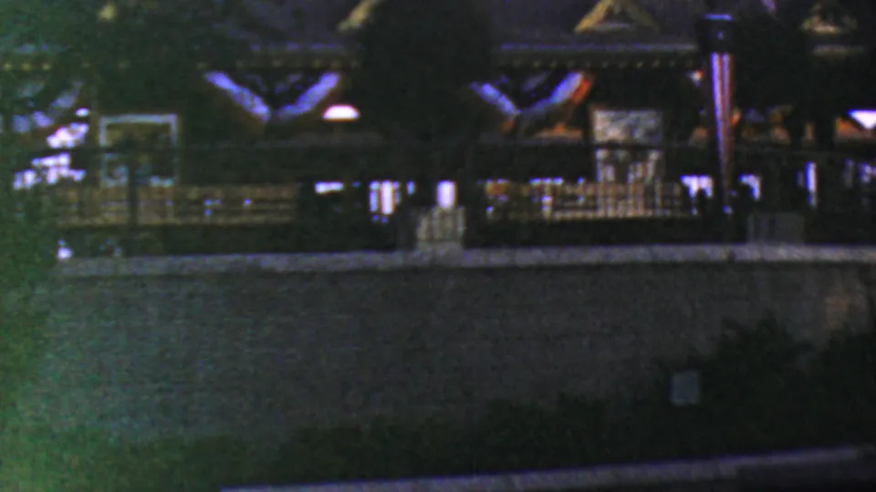 1964 Anaheim Disneyland home-movie frame showing a dark park structure with railings, low lamps, trees, and a foreground wall in soft night exposure.