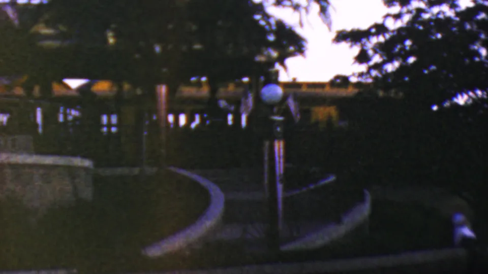 1964 Anaheim Disneyland home-movie frame looking across a landscaped night path with globe lamps, trees, railings, and distant park buildings.