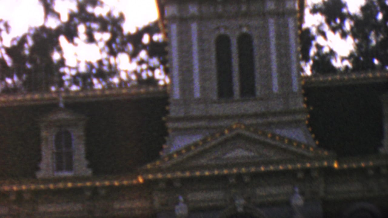1964 Anaheim home-movie frame showing the upper portion of Disneyland City Hall, with illuminated trim, arched windows, rooflines, and silhouetted trees in low light.