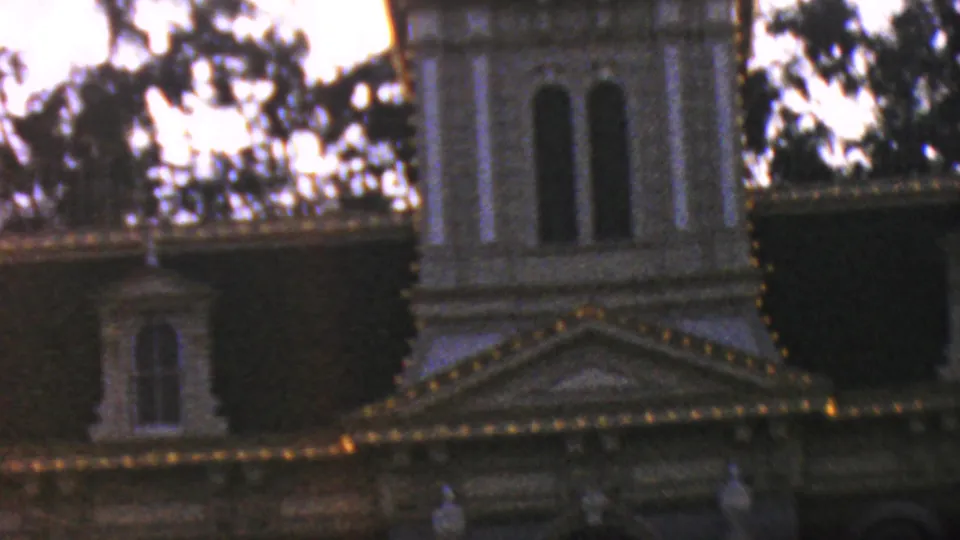 1964 Anaheim home-movie frame showing the upper portion of Disneyland City Hall, with illuminated trim, arched windows, rooflines, and silhouetted trees in low light.