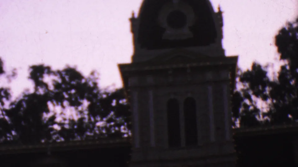 1964 Anaheim visitor footage frame with the darker tower, roof edge, arched openings, and tree silhouettes around the Disneyland City Hall building.
