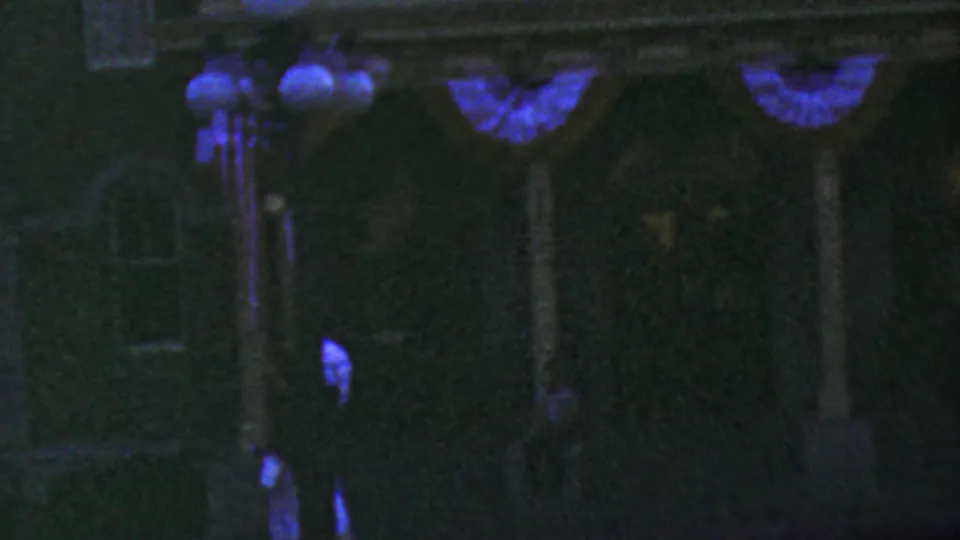1964 Anaheim home-movie frame showing a darker section of the Disneyland City Hall facade with bunting, globe lamps, doorway shapes, and indistinct shadowed visitors near the lower edge.