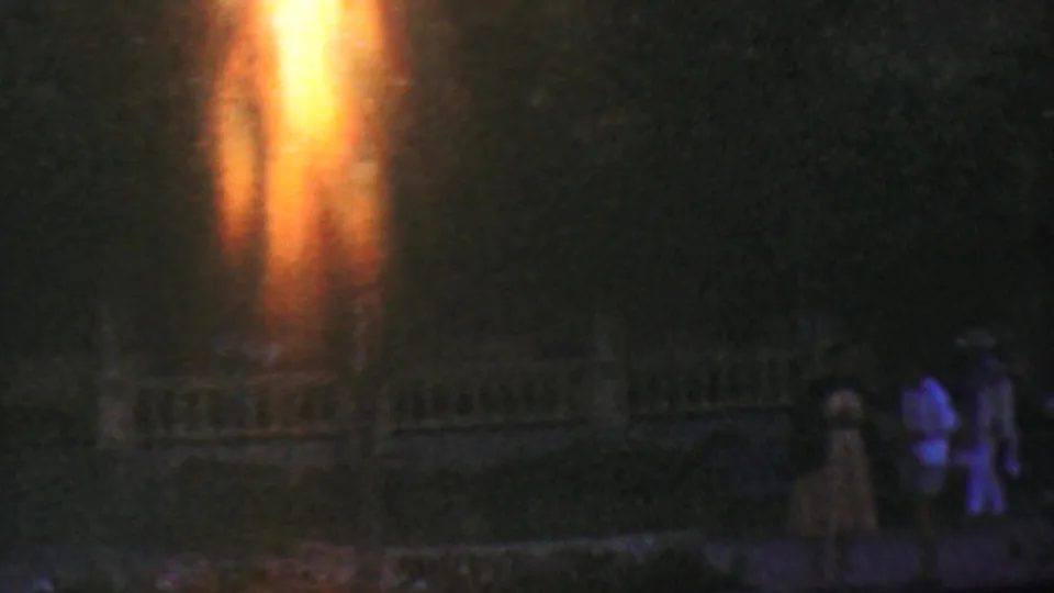 1964 Disneyland nighttime home-movie frame with visitors near a low fence or railing, a dark building behind them, and warm orange flare across the scene.