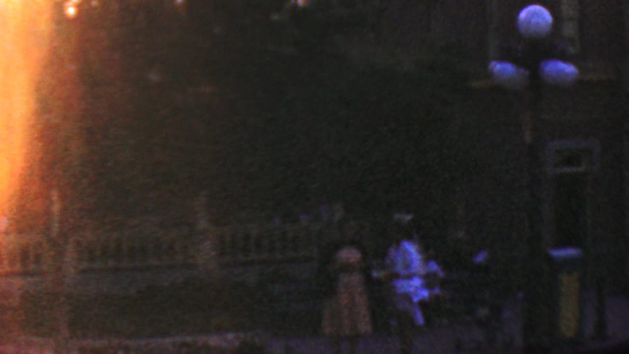 1964 Disneyland nighttime home-movie frame showing a dim walkway, silhouetted visitors, round exterior lamps, a building facade, and a strong orange light flare along the left edge.