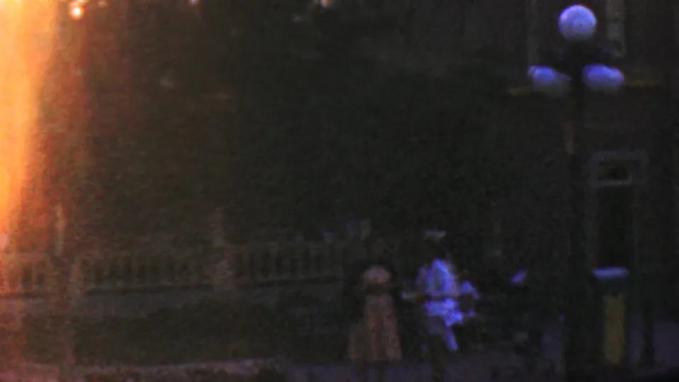 1964 Disneyland nighttime home-movie frame showing a dim walkway, silhouetted visitors, round exterior lamps, a building facade, and a strong orange light flare along the left edge.