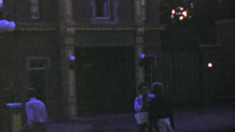 1964 Disneyland nighttime frame with shadowed visitors near a building front, round lamps overhead, a dark doorway, and soft blue-purple 8mm color cast.