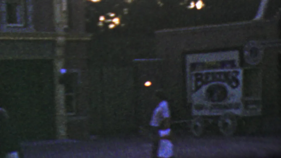 1964 Disneyland nighttime home-movie frame showing a dark storefront-like facade, a walking visitor, round lamps, and a partially legible illuminated sign that appears to include commercial lettering.