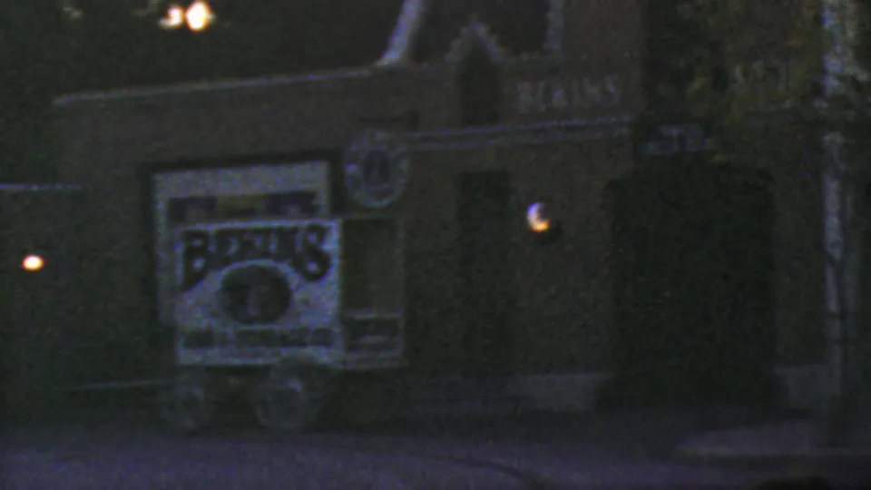 1964 Disneyland nighttime frame with a dark facade, partially readable illuminated commercial-style signage, small round lights, and dense tree shadow along the right side.