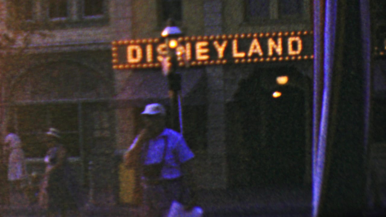Illuminated Disneyland sign over a dark entrance area with a man in a white hat walking near a lamppost.