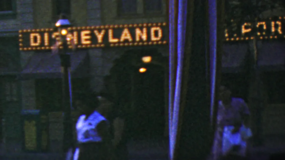 A low-light 8mm frame shows the Disneyland sign and entrance area with visitors passing below, partially obscured by vertical foreground poles.