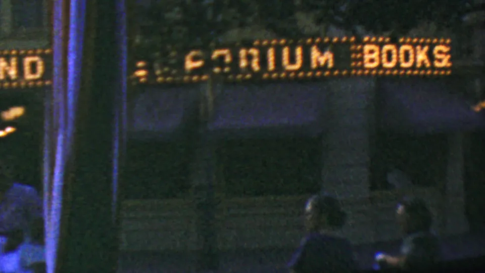 The Emporium Books marquee glows above a dim Disneyland storefront as visitors stand or walk in the lower foreground of the 1964 home-movie frame.